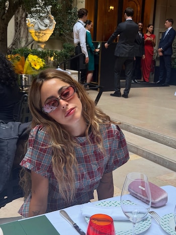 Mujer con cabello largo castaño, blusa a cuadros rojos y azules, y gafas de sol rosas, sentada en una mesa con un plato, cubiertos y dos copas