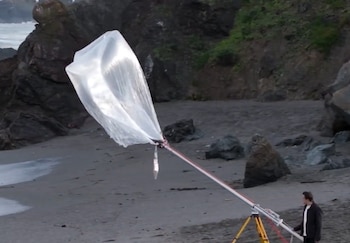 Un hombre junto a un tubo sobre un trípode con una bolsa de plástico inflada por el viento en una playa rocosa