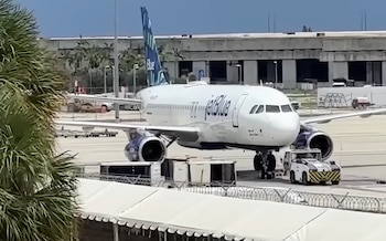Avión de JetBlue en pista de aeropuerto con vehículos de asistencia, edificios y palmeras al fondo. Barricada y tiendas blancas en primer plano