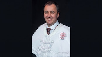 Hombre con camisa, corbata y muceta ceremonial blanca con el emblema de la PUCPR sobre un fondo negro.