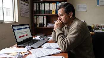 Hombre sentado frente a una laptop en un escritorio con papeles y calculadora; la pantalla muestra 'TRANSFERENCIA INTERNACIONAL PENDIENTE'.