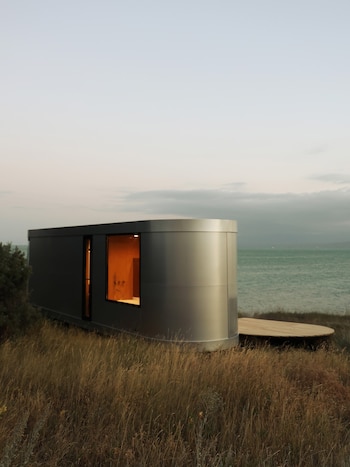 Estructura metálica de diseño moderno con ventana iluminada en un campo de pasto alto junto al mar