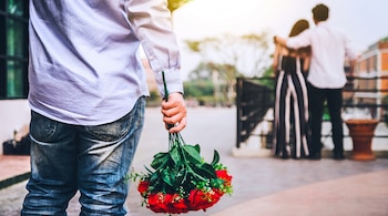 Hombre de espaldas con camisa azul y pantalón vaquero sostiene ramo de rosas rojas. Pareja abrazada al fondo, desenfocada