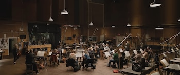Gran estudio de grabación musical con orquesta, atriles, instrumentos, micrófonos, monitores, luces de techo y una sala de control con personas