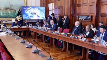 Personas con vestimenta formal sentadas ante una mesa con micrófonos y pantallas en una sala con paneles de madera y cuadros