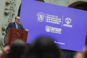 Un hombre de traje habla en un podio de madera junto a una pantalla azul con logotipos del Gobierno de Guatemala
