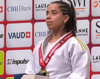 Una mujer con traje blanco de judo y medalla de oro al cuello posa frente a un fondo con logotipos