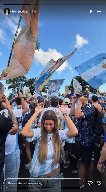 Mujer con camiseta argentina, jean, manos en alto, en medio de una multitud con camisetas de fútbol. Banderas de Argentina y de Diego Maradona. Cielo azul