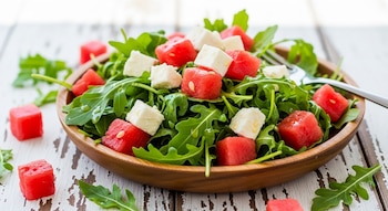 Cuenco de madera con ensalada de hojas verdes, cubos de sandía roja y cubos de queso blanco sobre una mesa rústica blanca con un tenedor.