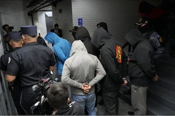 Agentes con chalecos tácticos y pasamontañas custodian a individuos con capuchas y esposas cerca de una reja metálica y vehículos