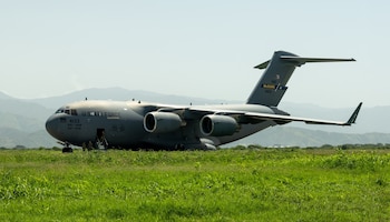 Avión grande militar gris con inscripción U.S. AIR FORCE, cuatro motores, puerta frontal abierta, personal militar, hierba y montañas distantes