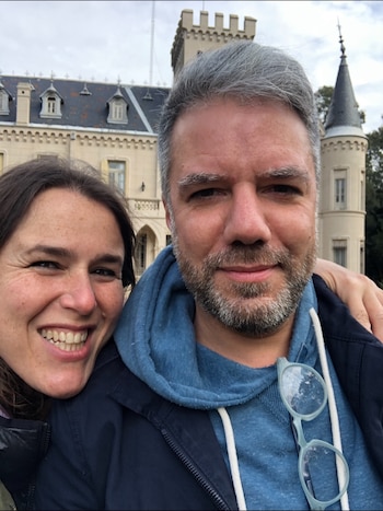 Primer plano de mujer con cabello castaño sonriendo junto a un hombre con cabello gris y barba, con anteojos en su sudadera. De fondo, un edificio antiguo con torreones