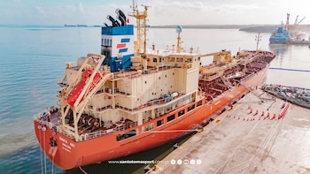 Buque tanque rojo y crema atracado en un muelle, con equipo de carga, balsas salvavidas y conos naranjas. Otro buque al fondo