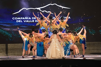 Diez bailarines actúan en un escenario. La mayoría usa vestuarios con alas doradas y telas azules; un bailarín usa un vestido crema. Chispas y una pantalla de fondo