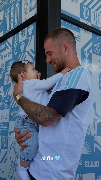 Alexis Mac Allister, sonriendo y con el pelo corto, sostiene en brazos a su hija Alaia, una bebé con un chupete rosado y un lazo azul claro en el pelo, ambos se miran