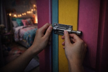 Dos manos cierran un cerrojo de metal en una puerta de paneles rosa, azul y amarillo. Al fondo se ve un dormitorio con cama y luces decorativas.