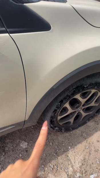 Una mano señala el neumático destrozado de un vehículo de color claro estacionado sobre un suelo de tierra