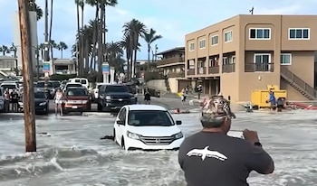 Un automóvil blanco avanza por una calle inundada frente a edificios, palmeras y personas que observan la escena