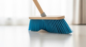 Una escoba nueva de mango de madera y cerdas de plástico azules empuja una pila de polvo en un suelo claro.