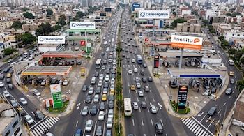 Vista aérea de una avenida con vehículos en marcha y estaciones de servicio con carteles de precios y paneles publicitarios a los lados.