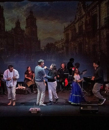 Varios artistas y músicos actúan y bailan en un escenario frente a un telón pintado con el paisaje de una ciudad colonial