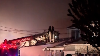 Un camión de bomberos con su escalera extendida hacia un techo, con dos bomberos en la cesta, mientras el humo se eleva sobre casas residenciales de noche