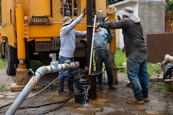 Tres hombres con sombrero operan una máquina de perforación junto a una tubería por la que sale agua hacia un balde.