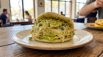 Arepa Reina Pepiada en primer plano sobre un plato blanco en una mesa de madera, rellena de aguacate, pollo y guisantes. Fondo de restaurante borroso con ventanas.
