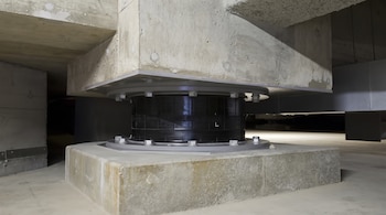 Un aislador sísmico de color negro y metal con pernos ancla una estructura superior de hormigón a una base de hormigón en un espacio interior.