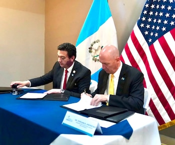 Dos hombres con traje firman y sellan documentos en una mesa. Detrás de ellos se observan las banderas de Guatemala y de Estados Unidos. Placa con el nombre Dr. Jorgan K. Andrews