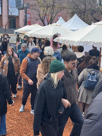 Rusherking y una mujer con gorro verde caminan entre personas en una feria al aire libre con sombrillas blancas