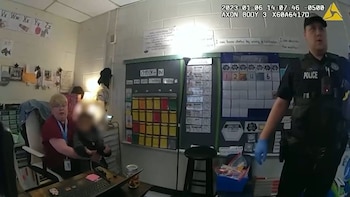 Un agente de policía permanece de pie en un aula escolar mientras una mujer sostiene en su regazo a un niño con el rostro borroso