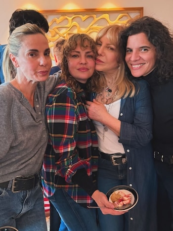 Cuatro mujeres posan juntas; una rubia, dos con cabello castaño y una con cola de caballo. Visten jeans y camisas informales. Una mujer sostiene un tazón con fresas