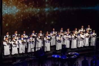 Un coro masculino con túnicas blancas y detalles dorados canta en un escenario iluminado junto a instrumentos de percusión