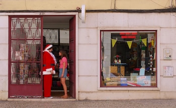Un hombre disfrazado de Santa Claus con barba blanca y traje rojo habla con una mujer de camiseta rosa en la entrada de una tienda con escaparate
