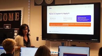 Una profesora habla frente a una pantalla digital interactiva con diapositivas en un aula con monitores de ordenador