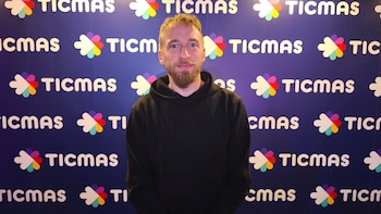 Hombre joven con cabello y barba castaños claros, vistiendo una sudadera negra, posa sonriendo frente a un fondo azul con logos de TICMAS