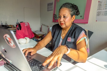 Una mujer latina de mediana edad con un chaleco de APPV trabaja en una computadora portátil plateada en un escritorio blanco, con auriculares al lado