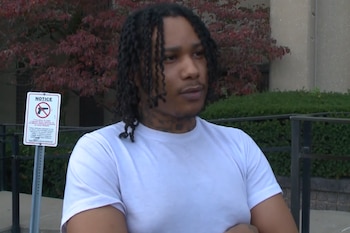 Un hombre joven con trenzas y camiseta blanca permanece de pie frente a un cartel de prohibición de armas y vegetación