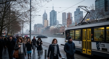 Una calle concurrida en Melbourne con personas caminando bajo un cielo nublado y un tranvía blanco y amarillo en movimiento. Se ven edificios altos y árboles sin hojas.