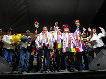 Un grupo de personas en un escenario durante un mitin político nocturno. Dos hombres, con ponchos y sombreros andinos coloridos, alzan el puño y un bastón festivo. Otros ondean