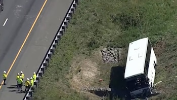 Vista aérea de un autobús blanco volcado en un terraplén junto a una autopista. Varios trabajadores con chalecos amarillos y cascos blancos están en el arcén