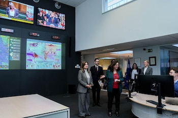 Una mujer de espaldas observa un muro con múltiples pantallas de televisión que muestran noticieros internacionales y mapas meteorológicos