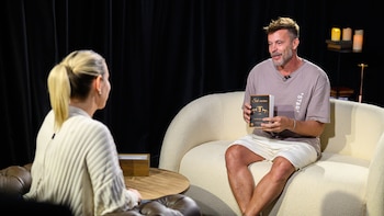 Alfredo Wiedmer, con camiseta gris y pantalones cortos claros, sonríe mientras sostiene su libro "Sub-marino" frente a una mujer rubia de espaldas