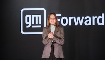 Una mujer con chaqueta marrón habla de pie frente a una pantalla negra con el logotipo gm y la palabra Forward