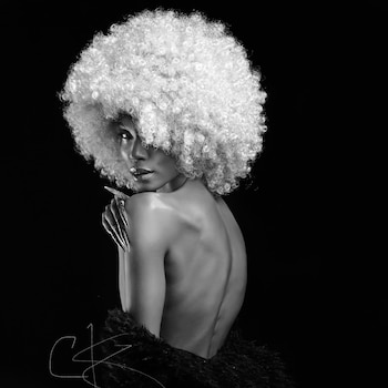 Mujer con peluca de cabello rizado blanco, espalda y hombros descubiertos, mano derecha con uñas largas y decoradas, sobre fondo negro. Fotografía en blanco y negro