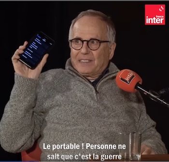 Hombre con gafas y jersey gris sostiene un smartphone negro en alto, hablando cerca de un micrófono rojo de France Inter. Fondo oscuro