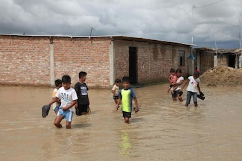 Un grupo de niños camina con los zapatos en la mano sobre agua acumulada frente a viviendas de ladrillo bajo un cielo nublado