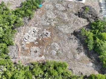 Fotografía aérea de un terreno despejado con estructuras de piedra circulares y cimientos, delimitado por árboles verdes