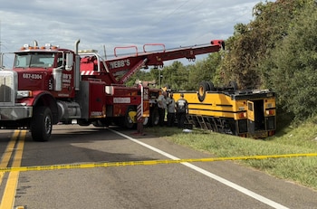 Una grúa roja de gran tamaño junto a un autobús escolar amarillo volcado al costado de una carretera acordonada con cinta de seguridad